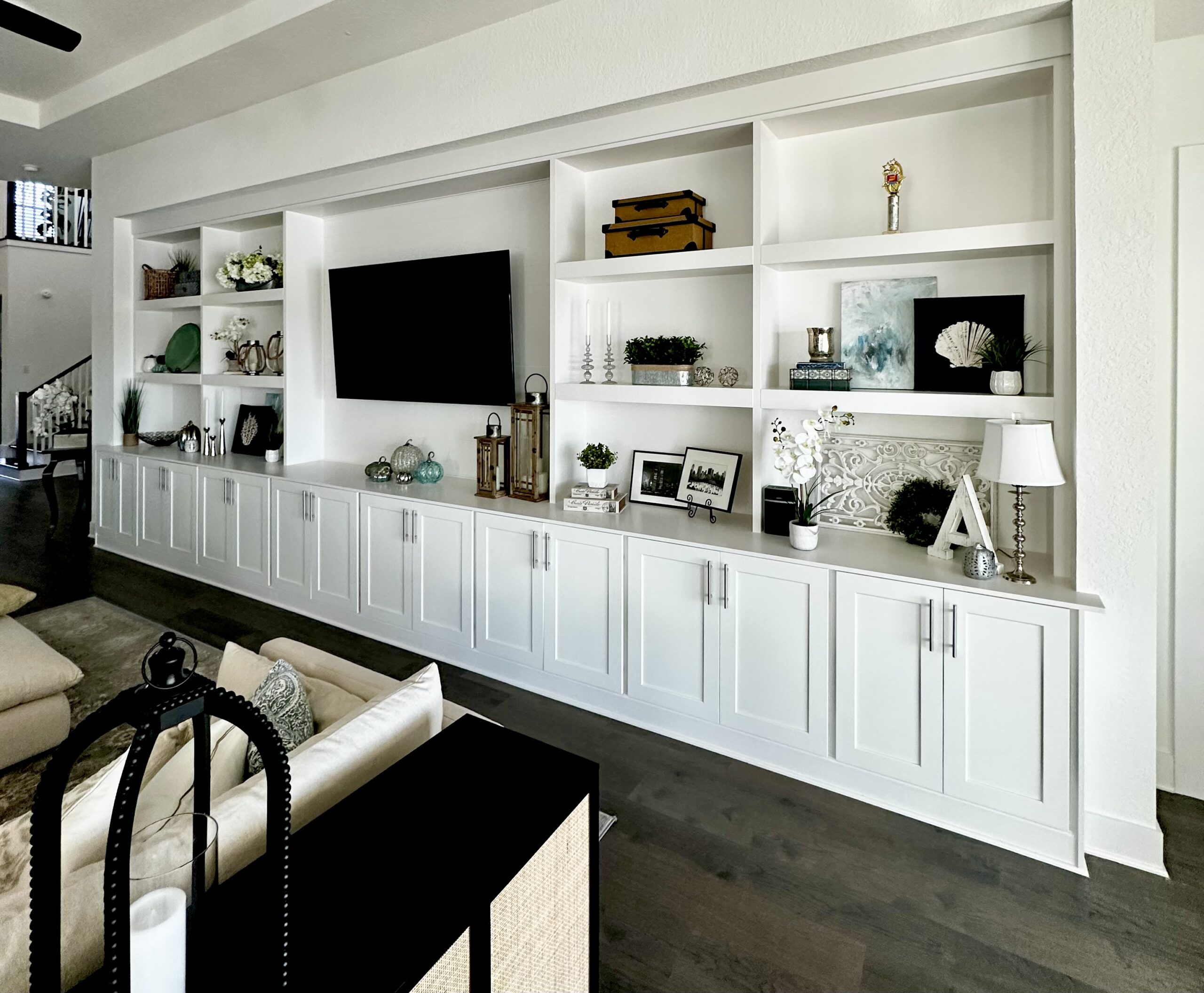 Living room wall featuring a custom white built-in unit with shaker-style lower cabinets and open upper cubby shelves displaying lanterns, vases, stacked boxes, and artwork, centered by a wall-mounted flat-screen TV above a wood countertop, set over dark hardwood flooring and beneath a tray ceiling.