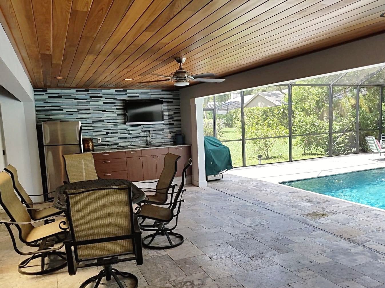 Outdoor covered patio with rich wood-plank ceiling featuring a ceiling fan, overlooking a dining table and pool area beyond a screened enclosure.