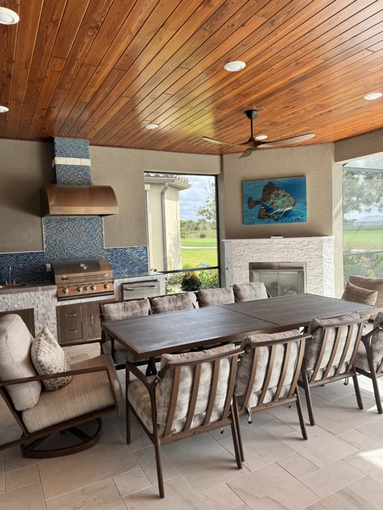 Covered patio featuring a rich wood-plank ceiling with recessed lights and a ceiling fan, a built-in stainless steel grill set into a white stacked-stone base with a colorful mosaic tile backsplash and vent hood, a white stacked-stone electric fireplace topped by a fish artwork, and a rectangular dining table surrounded by cushioned chairs on light tile flooring, overlooking a green lawn.