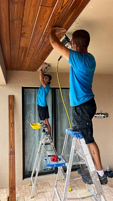 Two installers on aluminum ladders securing natural wood plank panels to a covered patio ceiling outside a stucco-clad home.
