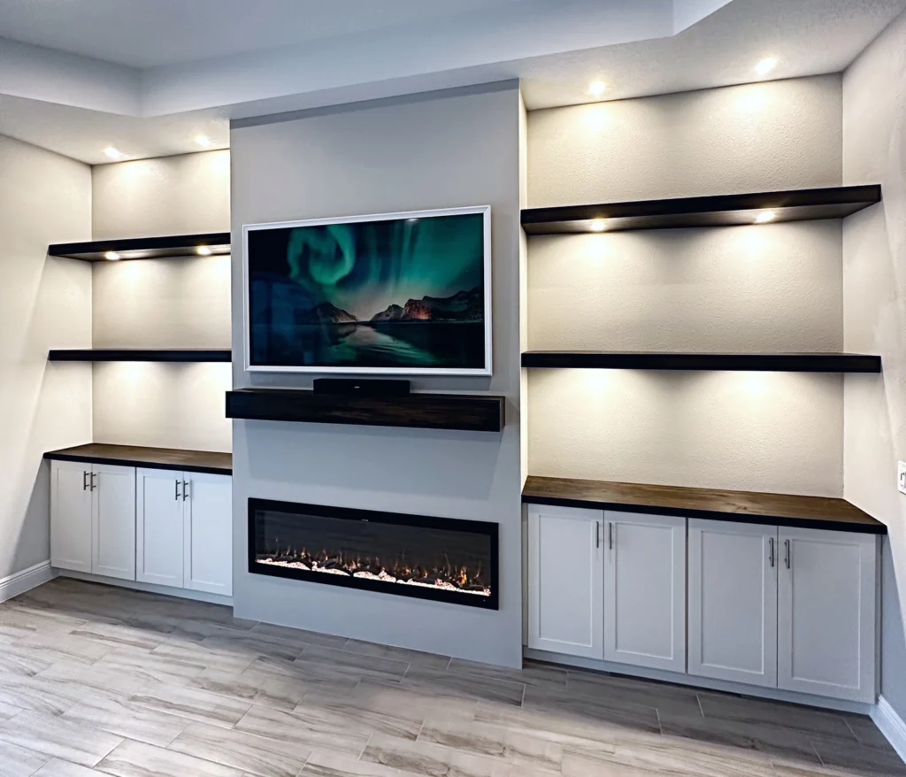 Gray feature wall with a wall-mounted TV and linear fireplace flanked by dark wood floating shelves and white cabinets, under a tray ceiling with recessed lighting.