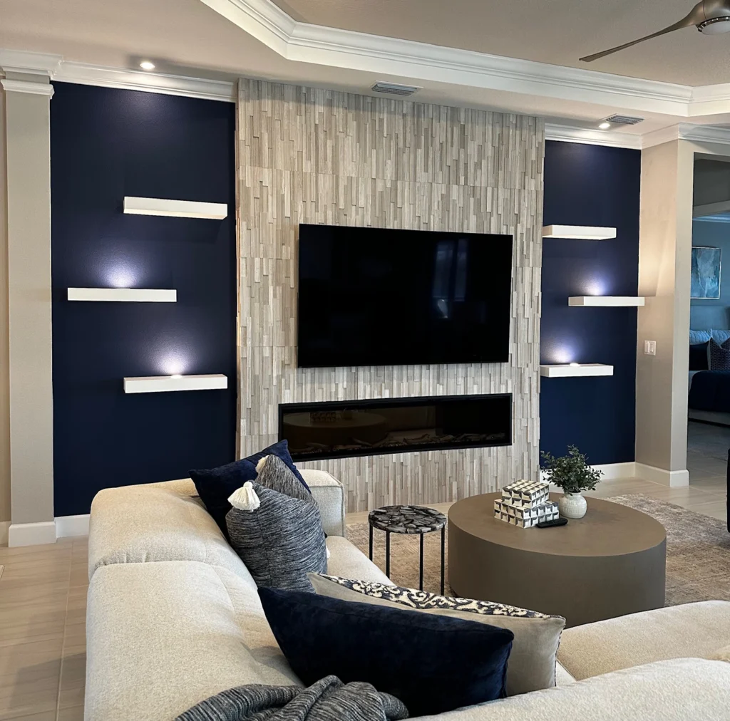 Central TV and linear fireplace set into a vertical wood mosaic tile panel, flanked by navy blue painted walls with white floating shelves and a tray ceiling.