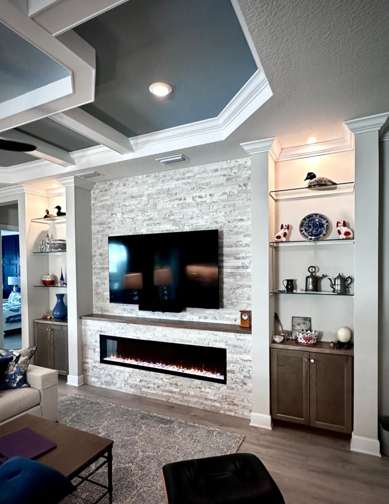 Floor-to-ceiling white stone veneer feature wall with a recessed display niche above a wall-mounted TV, a row of glass shelves on each side, a matching wood mantel, and a linear fireplace inset at the base.