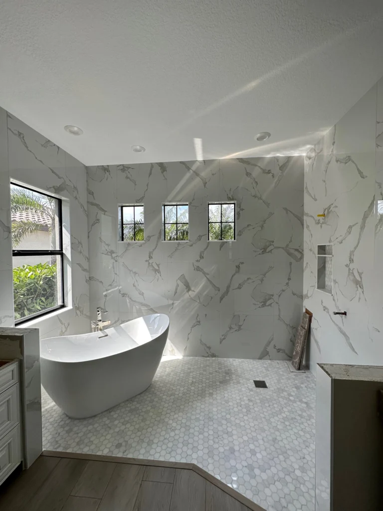 Spacious wet-room bathroom with a freestanding white soaking tub and open walk-in shower area, large-format marble-look floor and wall tiles, three small black-framed windows, recessed lights, and a built-in shower bench.