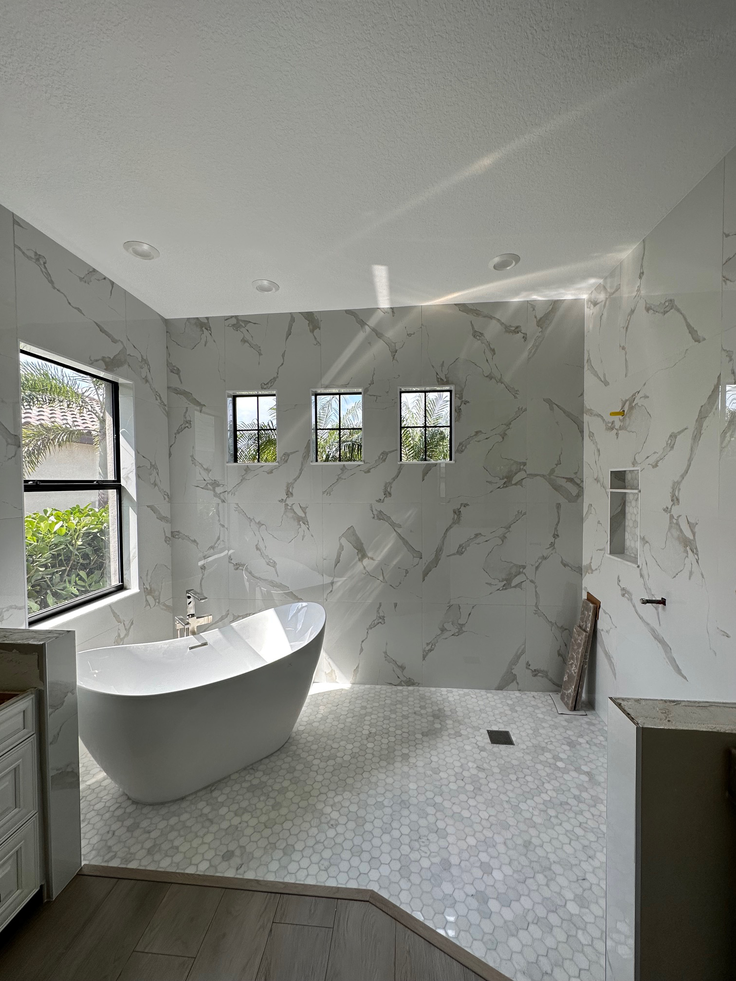 Spacious wet-room bathroom with a freestanding white soaking tub and open walk-in shower area, large-format marble-look floor and wall tiles, three small black-framed windows, recessed lights, and a built-in shower bench.