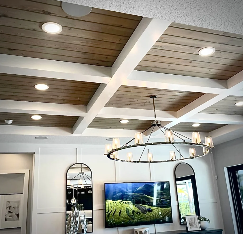 Ceiling with white-painted beams forming a coffered grid filled with natural wood plank panels and integrated recessed lights, centered by a circular metal chandelier with glass candle-style shades hanging above a living area.